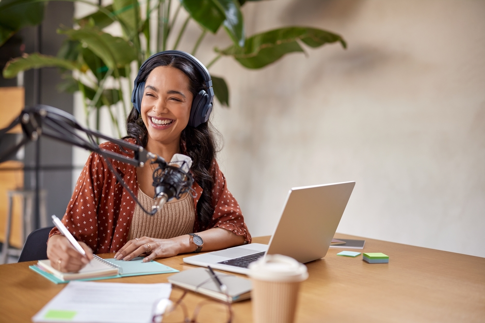 A podcaster recording an episode with an ad script, featuring a microphone, headphones, and a laptop.