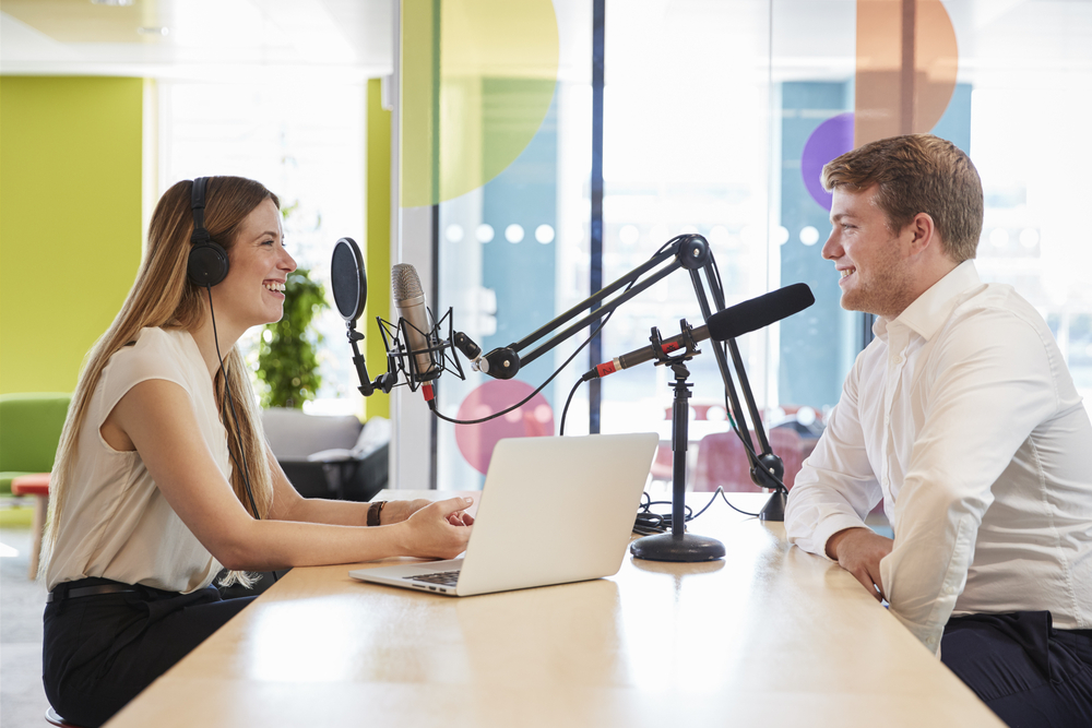 Young woman and man recording a podcast at a bright modern table with microphones and a laptop between them, smiling during conversation.