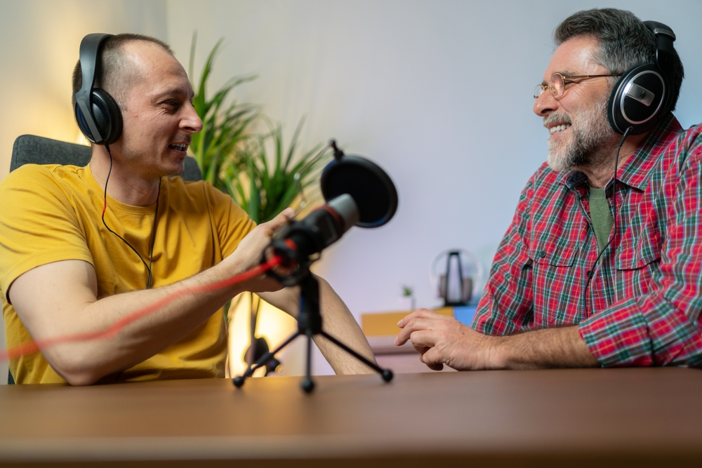 Two men wearing headphones laughing and talking during a podcast recording session at a desk with a plant in the background.
