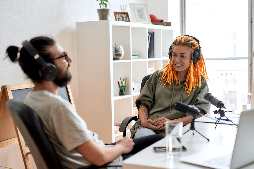 Young woman with orange dreadlocks and a green hoodie smiling during a podcast session with a male guest in a home studio setting.