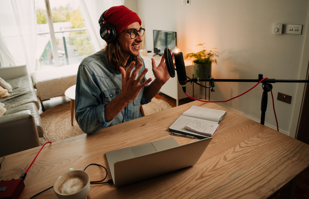 Man with long hair and red beanie passionately speaking into a microphone during a solo podcast recording in a cozy, modern apartment.